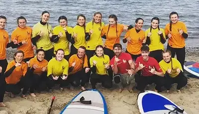 A group of people wearing yellow and orange shirts pose for a photo on a sandy beach near paddleboards by the water, smiling and making shaka hand signs.