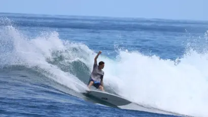 A surfer wearing a grey shirt and blue shorts rides a wave, carving through the water with his arm raised, surrounded by splashing surf on a sunny day in the sea.