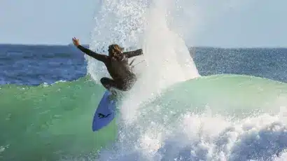 A surfer in a black wetsuit rides a turquoise wave, spraying water high into the air as they manoeuvre on a surfboard under a clear sky.