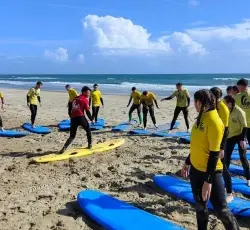 A group of people wearing yellow and red shirts stand on a sandy beach, practising surfing techniques on blue surfboards laid out on the sand, with the sea and a cloudy sky in the background.