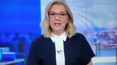 A woman with blonde hair and glasses, dressed in a dark top with a white collar, appears to be delivering news on television with a blue-themed newsroom background behind her.