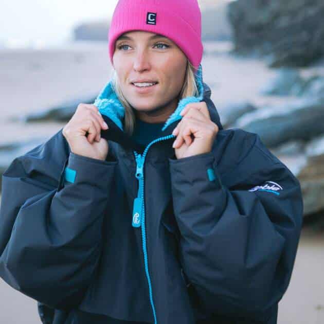 Wearing the C-Skins Storm Chaser 2mm Beanie in bright pink and a dark, oversized jacket, a woman stands on a rocky beach, glancing to the side as she adjusts her jacket collar.