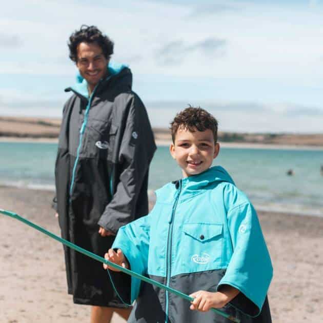 A smiling child in a blue Robie Dry-Series rain jacket holds a green fishing net on a sandy beach, with water and hills visible in the partly cloudy background. An adult in a dark rain jacket stands behind him.