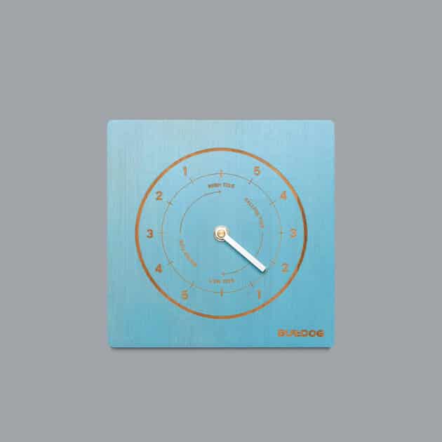 The Bulldog Single Dial Tide Clock is a square, blue clock with gold numbers and markings, white hands, and “SURFLOG” in gold at the lower right, displayed on a plain grey background.