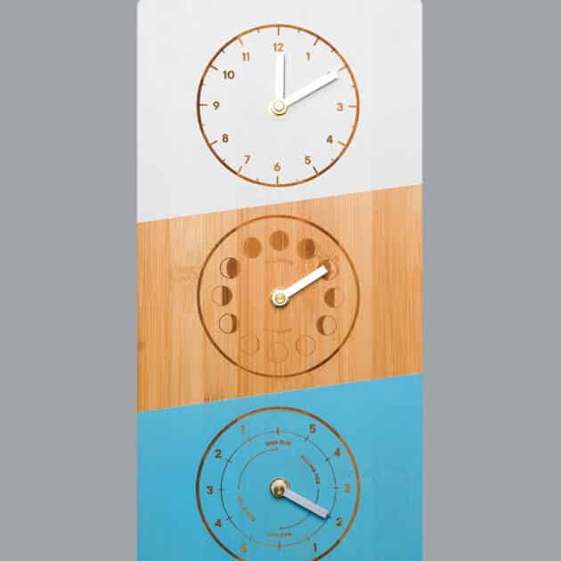 The Bulldog Triple Dial Tide Clock is a rectangular wall clock with three round dials—white, wood-coloured, and blue—on a grey background. Each dial has golden hands, unique markings, and "BULLDOG" in orange.