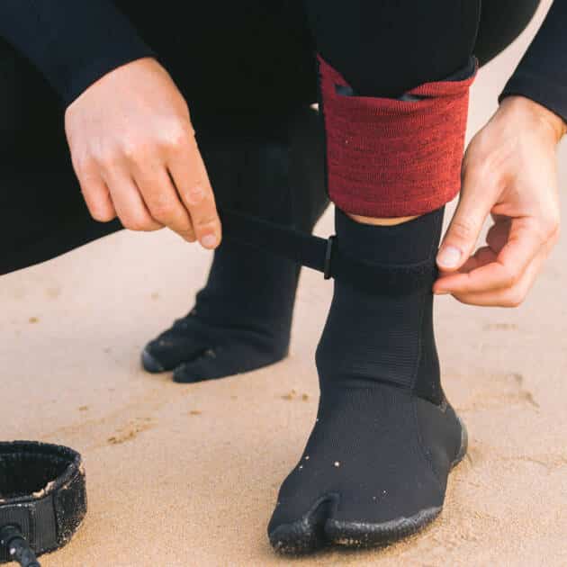 Wearing C-Skins Blackout 3mm Adult Split Toe Boots, a person in a wetsuit kneels on the sand, fastening the black surf boot with a strap. A red ankle band and a nearby surfboard leash complete the scene.