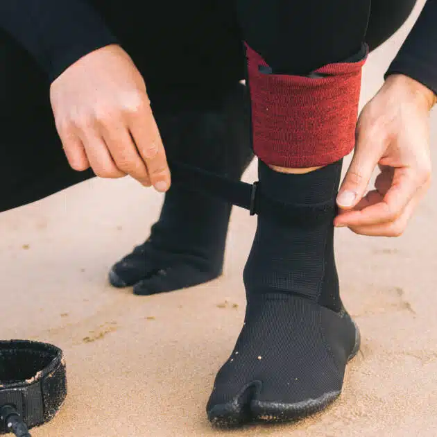 Wearing C-Skins Blackout 3mm Adult Split Toe Boots, a person in a wetsuit kneels on the sand, fastening the black surf boot with a strap. A red ankle band and a nearby surfboard leash complete the scene.