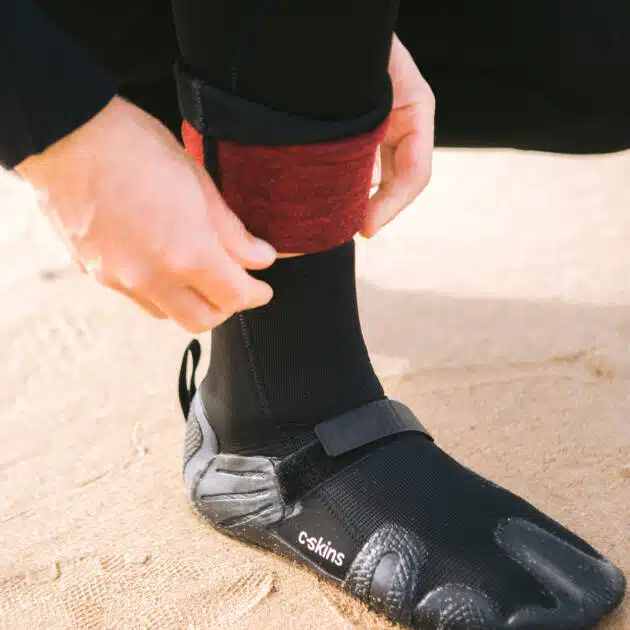 A person adjusts the red cuff of their wetsuit over C-Skins Wired 5mm Split Toe Boots with a strap and "c-skins" branding whilst standing on a sandy beach.