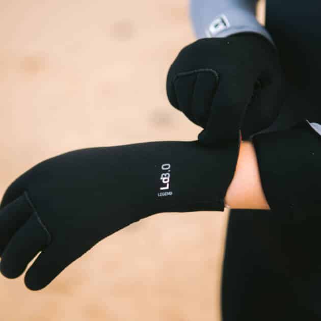 A person in a wetsuit pulls on C-Skins Legend 3mm Junior Gloves, featuring "LEGEND" markings on the wrist, as they get ready for water activities on a sandy beach.