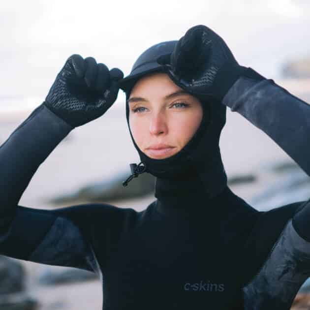 Wearing the C-Skins Legend 2.5 mm Hood and gloves, a person in a black wetsuit adjusts their hood whilst standing on a sandy, rocky beach.