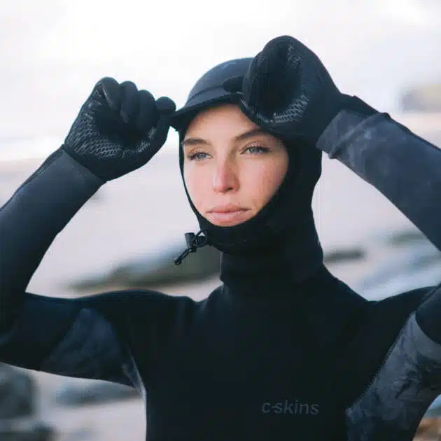 Wearing the C-Skins Legend 2.5 mm Hood and gloves, a person in a black wetsuit adjusts their hood whilst standing on a sandy, rocky beach.