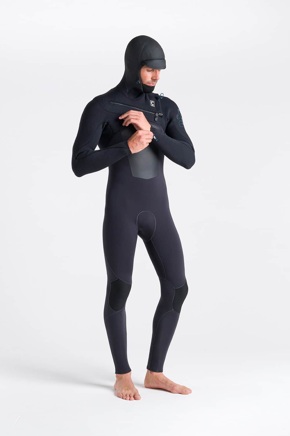 A man wearing the C-Skins Men's ReWired 5/4mm Hooded Wetsuit stands barefoot on a white background, adjusting the chest zip.