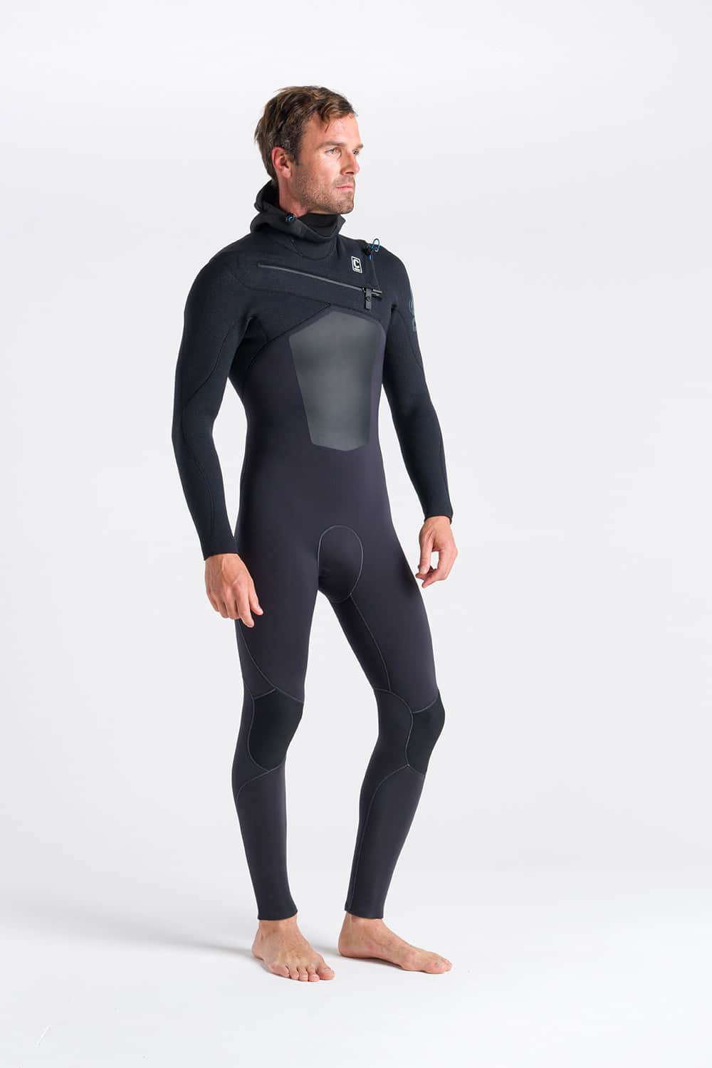 A man stands barefoot on a white background, wearing the C-Skins Men's ReWired 5/4mm Hooded Wetsuit. The black full-body wetsuit features a front zip and hood. He faces slightly to the side with his arms relaxed at his sides.