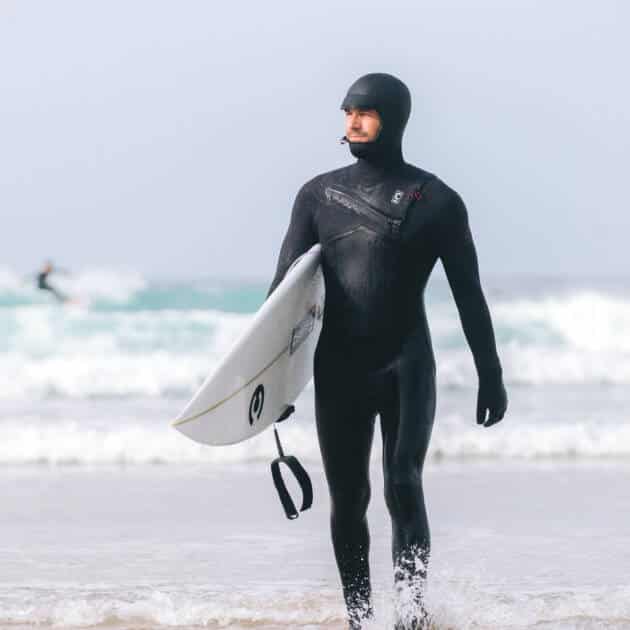 Wearing a C-Skins Men's ReWired 5/4mm Hooded Wetsuit, a surfer holds a white surfboard whilst walking from the sea onto a sandy beach, with waves and another surfer in the background.