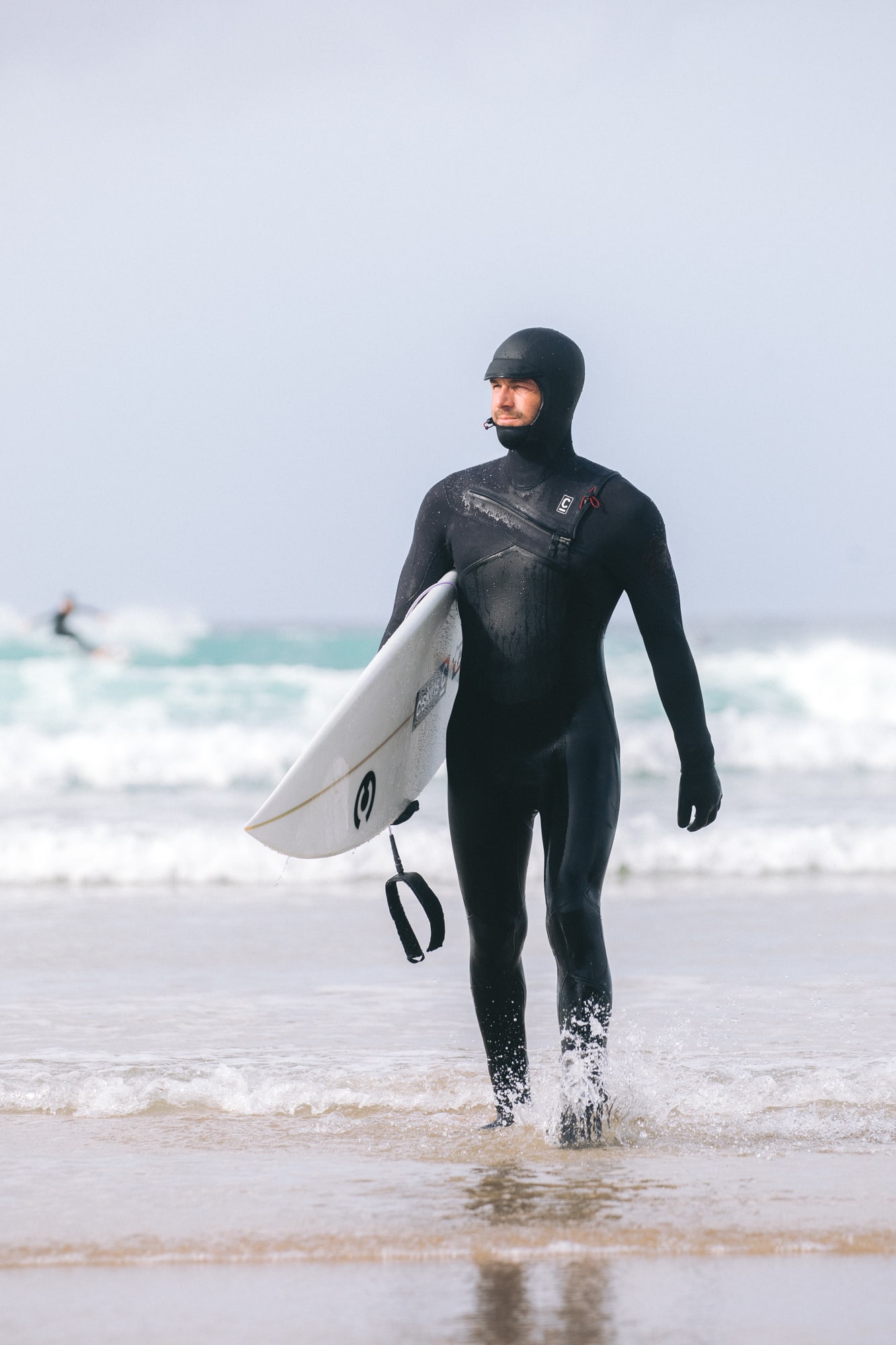 Wearing a C-Skins Men's ReWired 5/4mm Hooded Wetsuit, a surfer holds a white surfboard whilst walking from the sea onto a sandy beach, with waves and another surfer in the background.