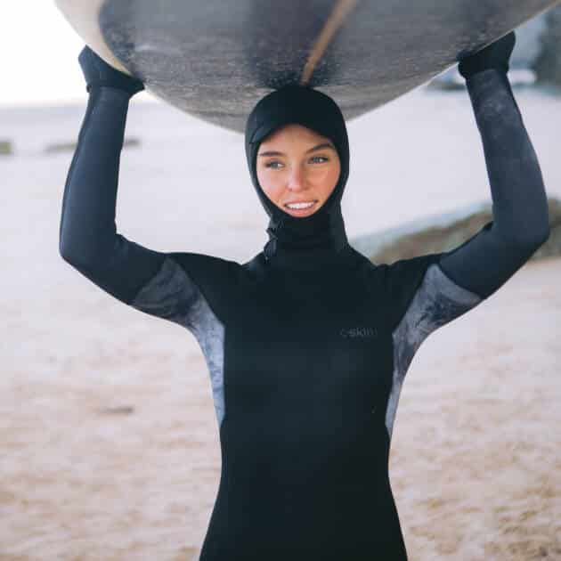 Wearing the Cskins Women’s Surflite 5/4/3mm Back Zip Wetsuit, a woman stands on a sandy beach, smiling at the camera whilst holding her surfboard above her head.