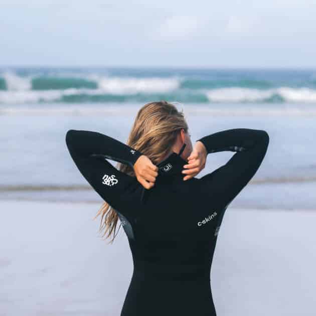 A woman with long hair stands on a sandy beach facing the sea, adjusting the zip of her C-Skins Women's Solace 4/3mm Chest Zip Wetsuit as waves roll in under a cloudy sky.