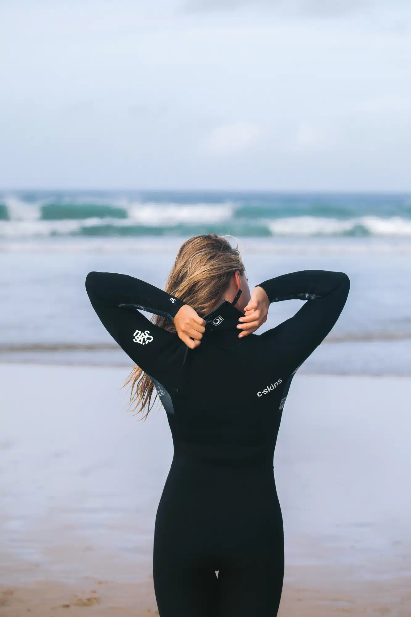 A woman with long hair stands on a sandy beach facing the sea, adjusting the zip of her C-Skins Women's Solace 4/3mm Chest Zip Wetsuit as waves roll in under a cloudy sky.