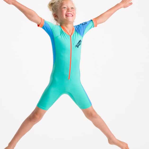 A smiling young child with blond hair in a ponytail jumps with arms and legs spread wide, wearing the C-Skins NuWave UV Sunsuit Shortie against a plain white background.