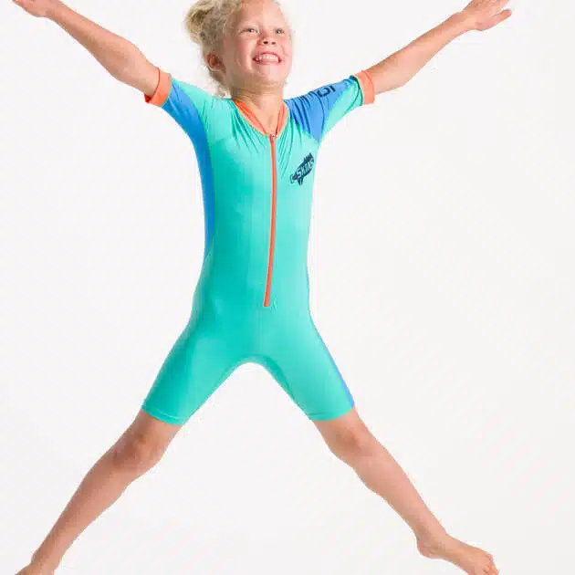 A smiling young child with blond hair in a ponytail jumps with arms and legs spread wide, wearing the C-Skins NuWave UV Sunsuit Shortie against a plain white background.