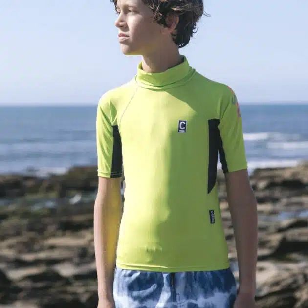 A boy in a C-Skins NuWave Rash X SS Vest and blue tie-dye swim shorts stands on rocky shore, gazing sideways with the sea and clear sky behind him.