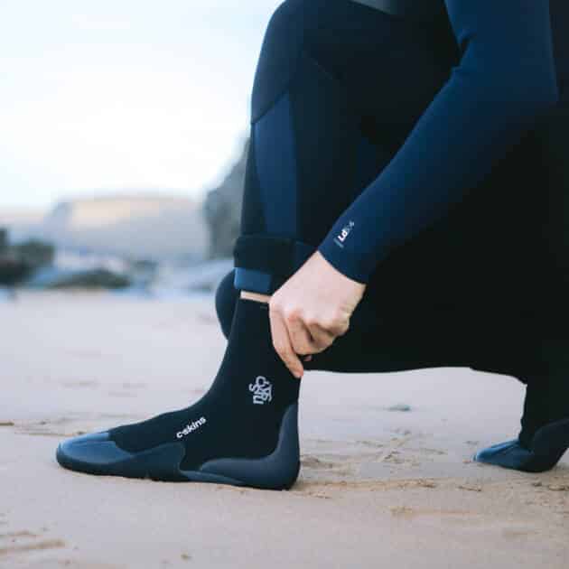 A person in a navy wetsuit kneels on a sandy beach, adjusting their cuff while wearing C-Skins Adult Legend 5mm Round Toe Boots. Their face is out of frame. There are rocks and water in the background.