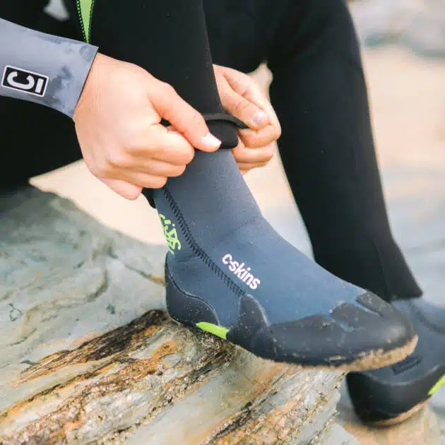 A person in a wetsuit sits on a rock and adjusts the ankle strap of the C-Skins Junior Legend 3.5mm Zipped Round Toe Boots, featuring grey and black neoprene with green accents, whilst sand covers the boot and surrounding rocks.