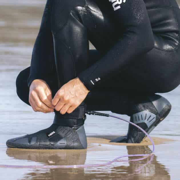 A person wearing C-Skins Session 7mm Round Toe Boots and a black wetsuit crouches on wet sand, attaching a surfboard leash to their ankle. Only their lower body and hands are visible, reflected in the shiny surface.