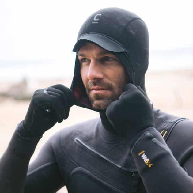 A man stands on a beach adjusting his C-Skins Wired 2mm hood, wearing a matching black wetsuit. The background is blurred, with sand and indistinct objects visible.