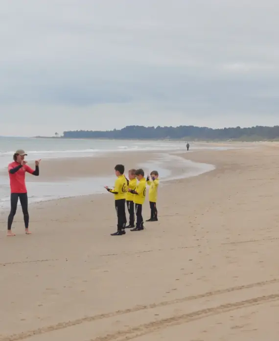 A person in a red jacket takes photos of four children wearing yellow jackets standing in a row on a wide, empty beach with gentle waves and a cloudy sky.