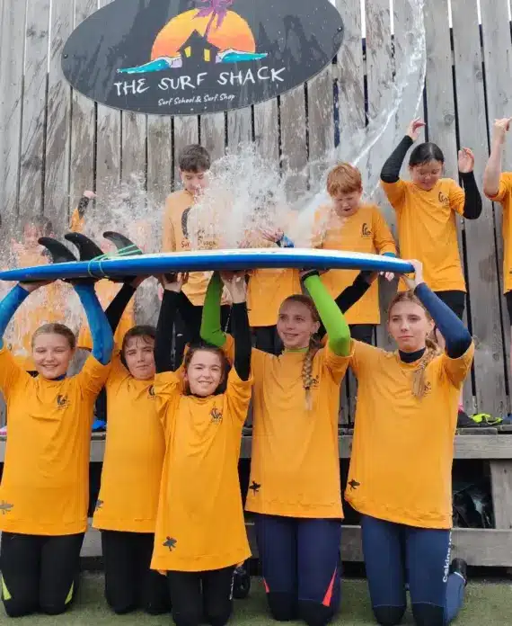 A group of kids in yellow surf shirts pose in front of a wooden fence with a "The Surf Shack" sign. Five kids hold a surfboard above their heads as water splashes onto them, whilst others cheer and smile in the background.