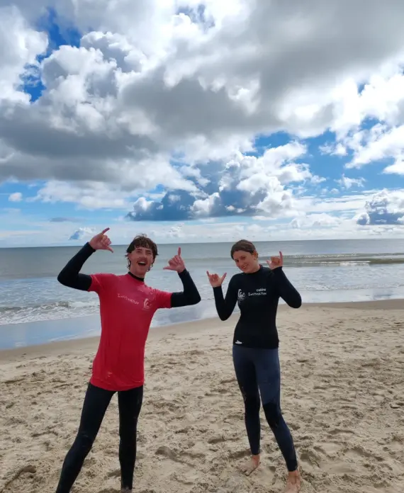 Two people in wetsuits stand on a sandy beach, smiling and making shaka hand signs. The sky is partly cloudy with patches of blue, and the calm sea is visible in the background.