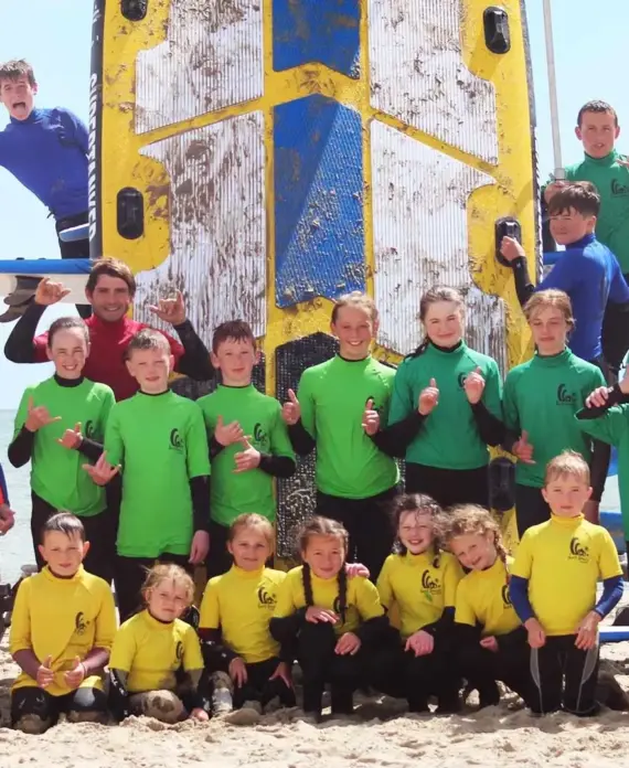 A large group of children in yellow and green shirts and a few adults in blue and red shirts pose and smile in front of a vertical surfboard on a sandy beach. Most give a thumbs-up gesture.