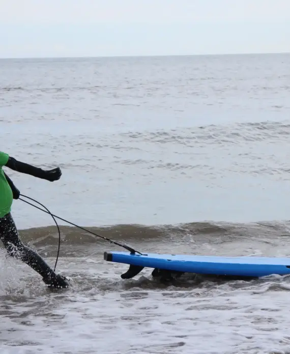A person in a green shirt and black wetsuit pulls a blue surfboard through shallow sea water, creating splashes as they walk towards the shore. The sea appears calm under a cloudy sky.