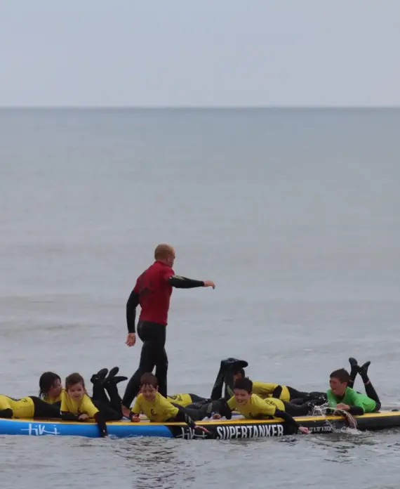 A group of children in yellow wetsuits lie on two large paddleboards in the sea, while an adult in a red wetsuit stands and gestures on one of the boards. The sky is overcast and the water is calm.
