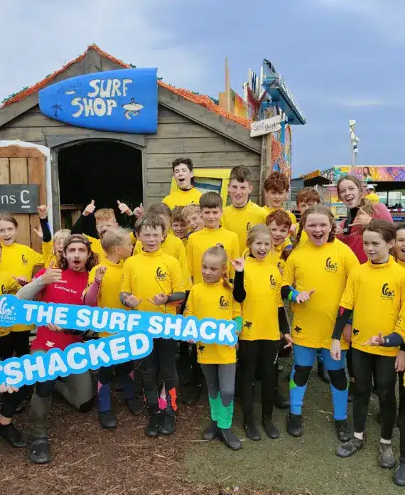 A group of smiling children and a few adults in bright yellow shirts pose excitedly outside a wooden surf shop, holding signs that read "The Surf Shack" and "Get Shack'ed." Surfboards and wetsuits are visible.