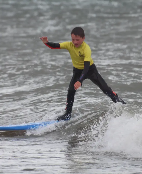 A young boy in a yellow shirt and black wetsuit rides a blue surfboard on a small wave in the sea, balancing with his arms outstretched.