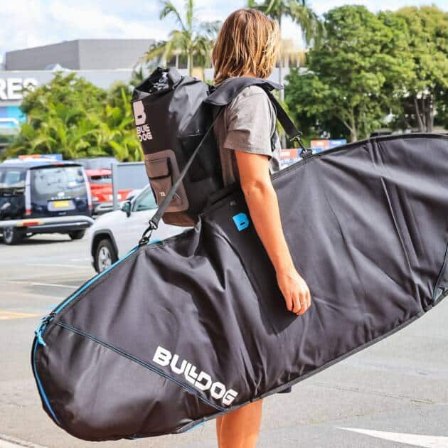 A person with a large black Bulldog Board Bag Core 5mm Fish and a rucksack stands in an airport departures car park, cars and trees behind them.