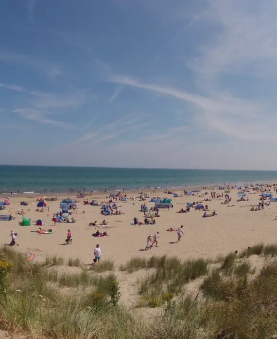 A wide sandy beach with people sunbathing, walking, and playing under a blue sky with light clouds. Colourful umbrellas and tents are scattered across the shore, with grass-covered dunes in the foreground.