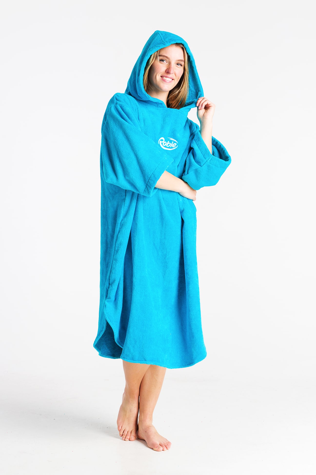 A person stands barefoot against a white background, smiling and wearing a loose-fitting, bright blue Robie Long Sleeve Towel Robe with a logo on the chest.