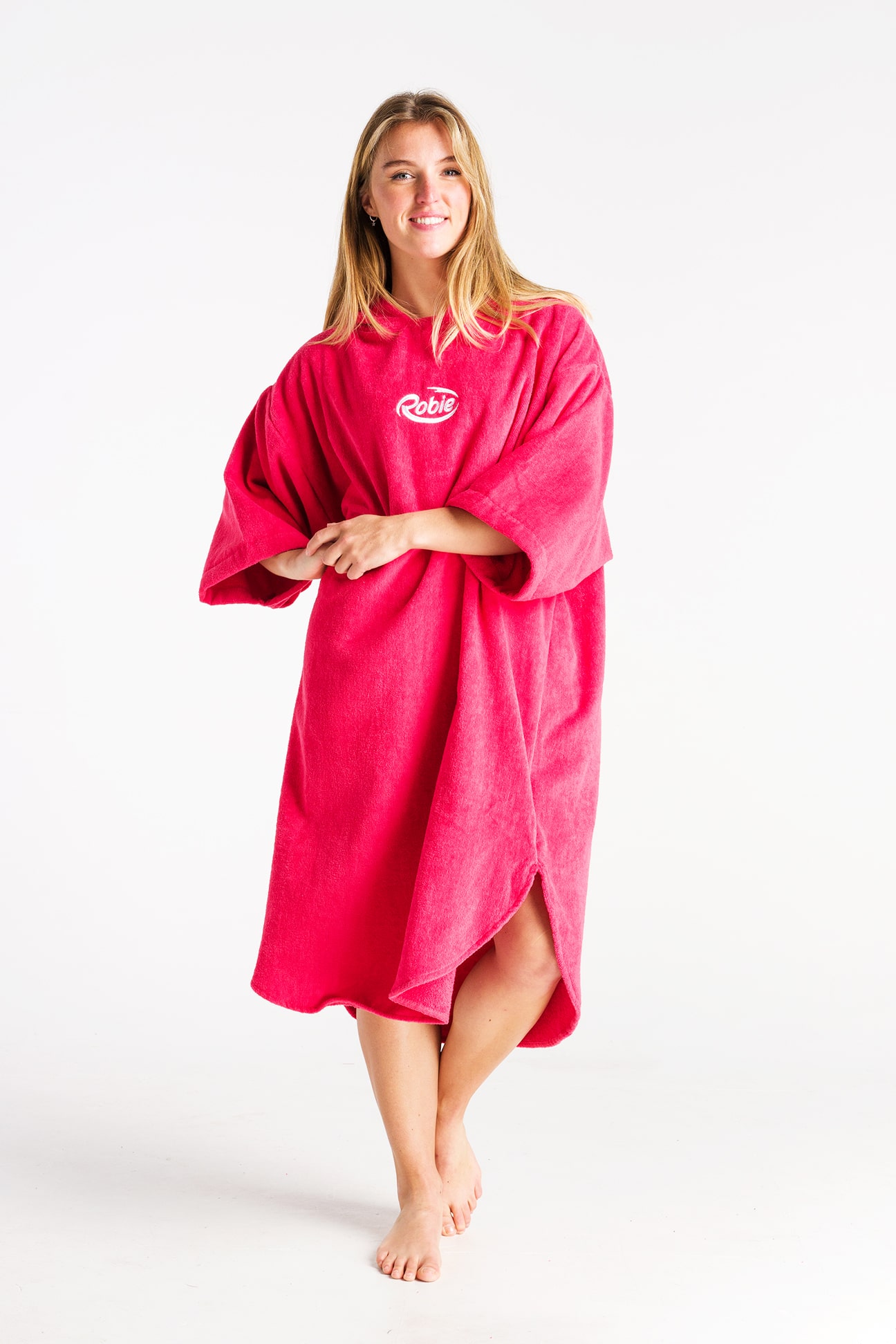 A person stands barefoot against a white background, smiling in a bright pink Robie Long Sleeve Towel Robe with "Robie" on the chest and wide sleeves, arms relaxed.
