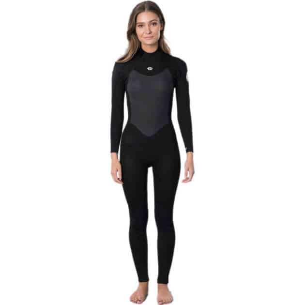 A woman stands facing forwards in the Ripcurls Women's Omega 4/3mm Back Zip Wetsuit, a full-length black suit with long sleeves and a lighter chest panel. She is barefoot, smiling slightly, and has long hair worn down.