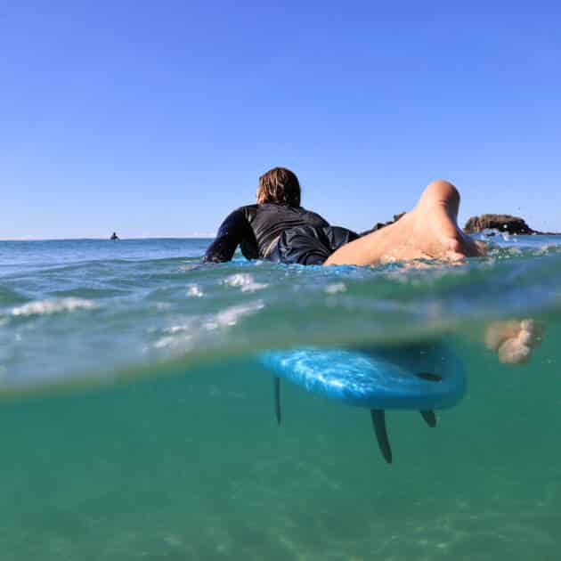 Wearing a black wetsuit, a person paddles on a Vision Surfboard Ignite across clear sea water beneath a bright blue sky, heading towards gentle waves in the distance.