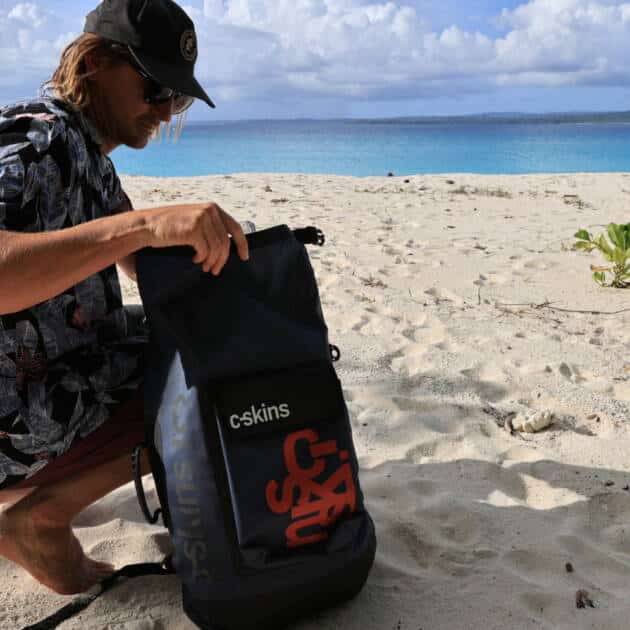 Wearing a black cap and floral shirt, a person kneels on sandy beach near the sea, looking into a C-Skins Storm Chaser Dry Bag beneath a partly cloudy sky.