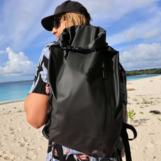 A person wearing sunglasses and a tropical shirt stands on a sandy beach with clear blue water, carrying the Cskins Session Dry Bag. Green plants and a partly cloudy sky are visible in the background.
