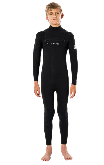 A young boy stands barefoot facing forwards, wearing the Ripcurl Junior Dawn Patrol 5/3mm Chest Zip Wetsuit—a full-length black suit with long sleeves and small logos on the chest and arm.