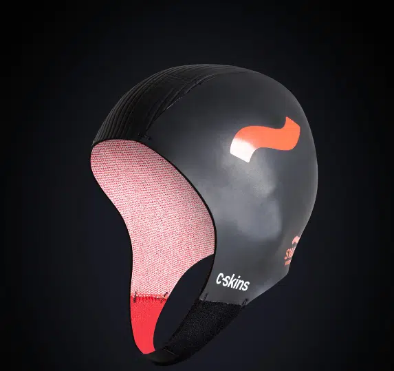 The C-Skins Swim Research 3mm GBS Cap is a black neoprene swim cap with a chin strap, red inner lining, and an orange wavy logo, shown against a dark gradient background.