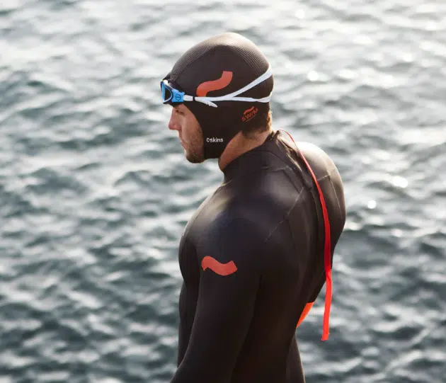 A swimmer in a black wetsuit and C-Skins Swim Research 3mm GBS Cap with orange markings stands by the water, wearing goggles and a cap with a red strap, looking out over the surface.