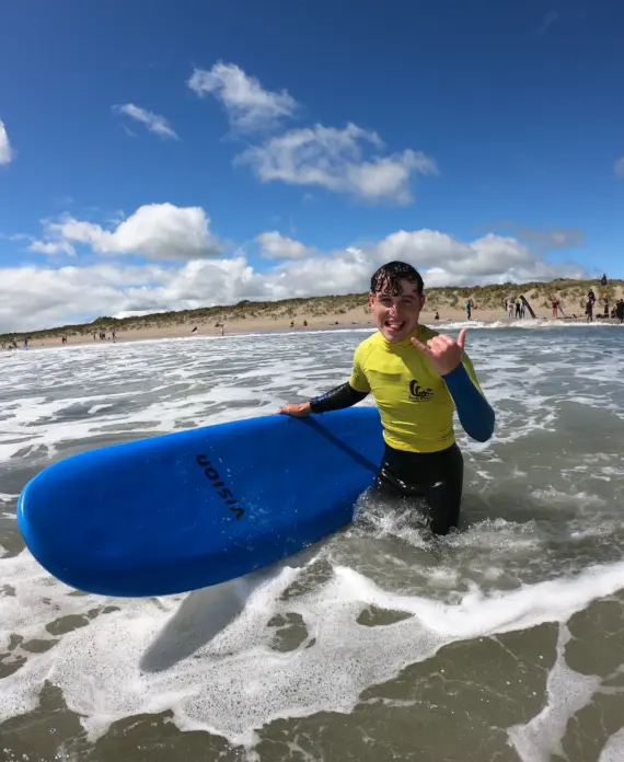A smiling person in a yellow rash vest stands in shallow sea water holding a blue surfboard, making a “shaka” hand sign. Sandy beach and people are visible in the background under a blue sky with clouds.
