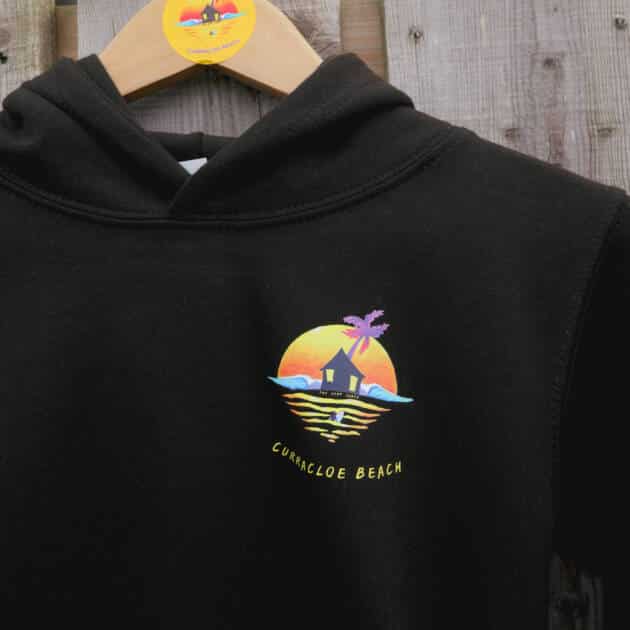 Shack Brand Curracloe Beach Kids Hoodie in black, displayed on a wooden hanger with a matching round tag, features a colourful sunset graphic with hut, palm tree, waves, and "Curracloe Beach" text.